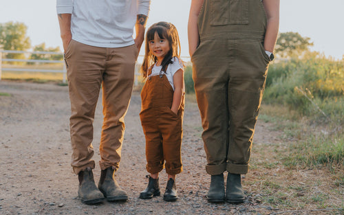 A young girl stands between two adults on a gravel path, all wearing brown pants and boots. The adults faces are not visible. The girl smiles, and the scene is outdoors in warm, golden sunlight.