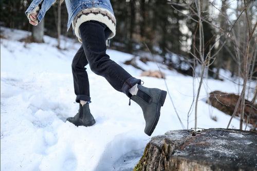 A person wearing black boots, dark pants, and a denim jacket steps onto a snow-covered tree stump in a snowy forest. Only the lower half of their body is visible.