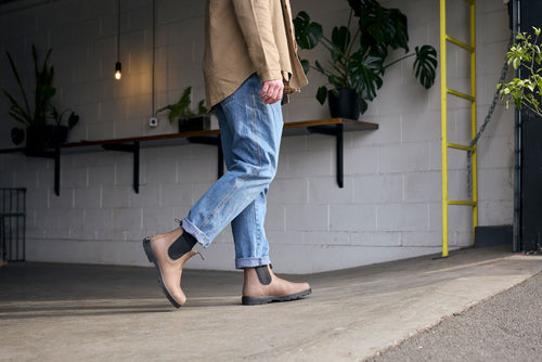 A person wearing rolled-up blue jeans and tan Chelsea boots walks indoors near a gray wall with plants on shelves and a yellow ladder visible in the background.