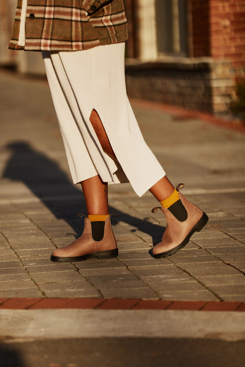 A person wearing a white ribbed skirt with a side slit, brown boots, and yellow socks walks on a sunlit sidewalk. The image is cropped above the knees and part of a plaid coat is visible.