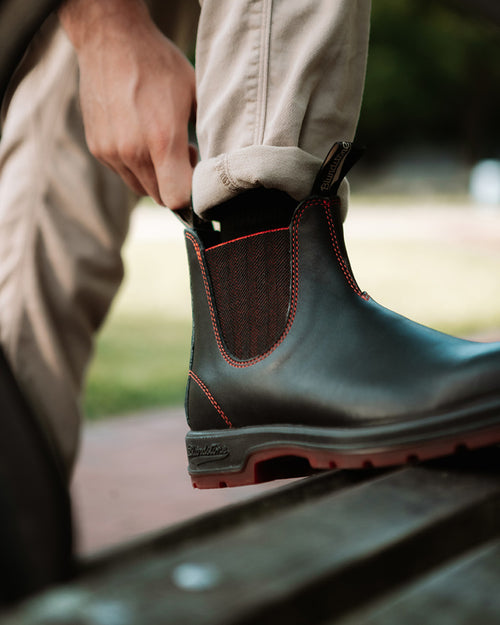 A person pulls on a black ankle boot with red stitching while wearing light-colored pants, sitting on a wooden bench outdoors.