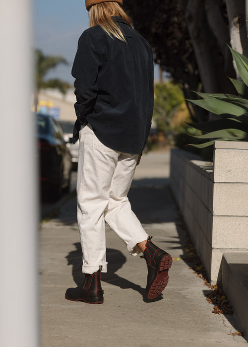A person with long hair walks on a sidewalk, wearing a black shirt, white pants, brown ankle boots, and a tan beanie. Their back is to the camera, and they are near plants and parked cars.
