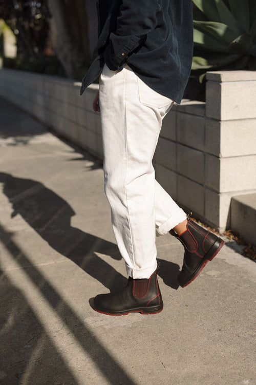A person wearing white pants, a dark jacket, and black boots with red stitching walks on a sidewalk next to a low concrete wall and green plants in sunlight.