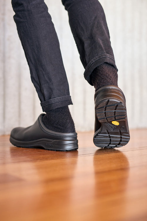 A person wearing black pants, black socks, and black slip-on shoes is walking on a wooden floor, viewed from behind at ankle level. The shoe soles are visible.