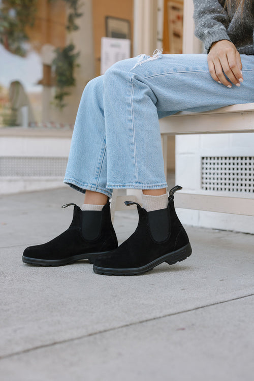 A person wearing light blue jeans and black Chelsea boots sits on a white bench outside, with one hand resting on their knee and part of their gray sweater visible.