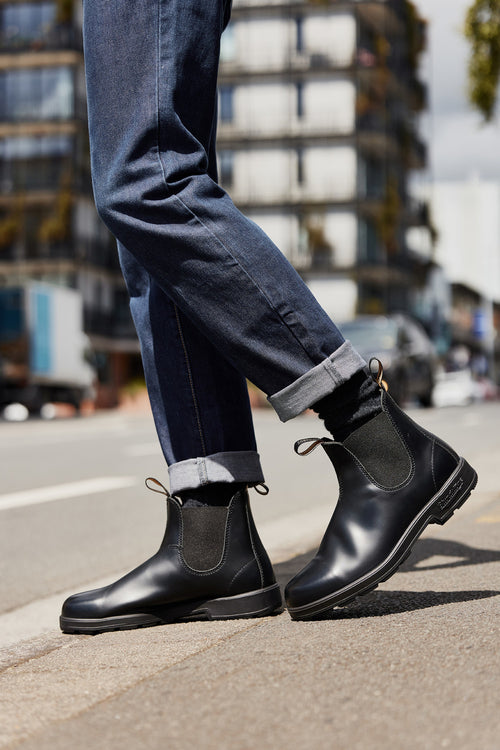 A person wearing cuffed blue jeans and black Chelsea boots stands on a city sidewalk, with blurred urban buildings and parked cars in the background.