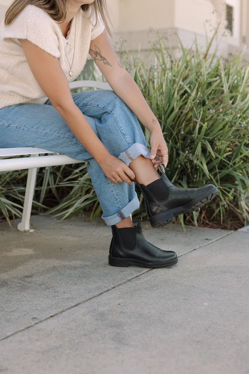 A person sitting on a white bench outdoors pulls on black ankle boots over rolled-up blue jeans, next to tall green plants. The person wears a short-sleeve top and has a tattoo on their left arm.