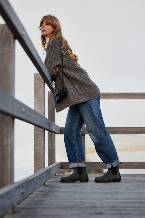 A woman with long hair leans on a wooden railing, wearing a gray sweater, blue jeans, and green boots. She holds a camera and looks thoughtfully into the distance against an overcast sky.