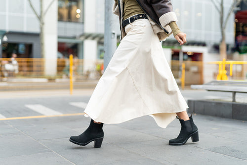 A person wearing a long cream skirt, black heeled boots, and a brown jacket walks on a city sidewalk, with blurred buildings and a street in the background.
