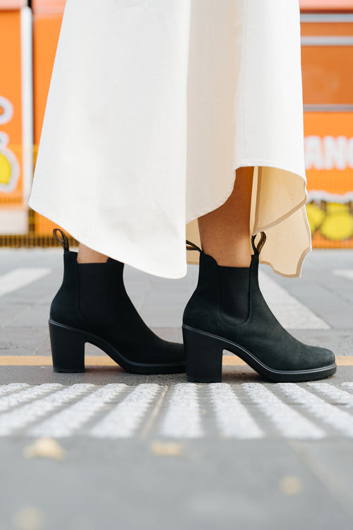 A person wearing a long white skirt and black heeled ankle boots stands on a crosswalk with an orange background.