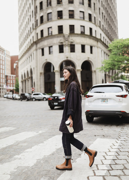 A woman wearing a dark oversized jacket, rolled-up jeans, and tan boots walks across a cobblestone street in a city, holding a coffee cup. A tall, curved building and several cars are in the background.
