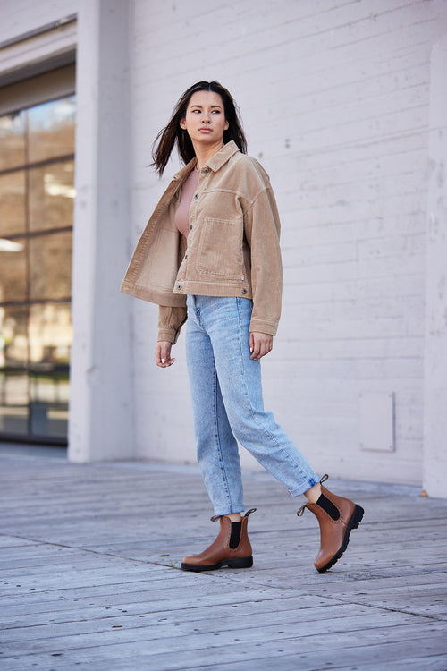A woman stands outdoors on a wooden deck, wearing a tan jacket, light blue cuffed jeans, brown ankle boots, and a light pink top. She looks to the side with one arm swinging naturally as she walks.