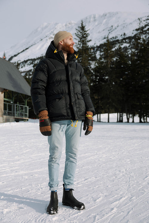 A bearded man in a black puffer jacket, beige knit hat, gloves, light jeans, and black boots stands on snow, with trees and a snow-covered mountain in the background.