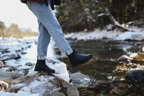 A person wearing cuffed jeans and black boots balances on a rock beside a shallow, snow-lined river in a forested area during winter.