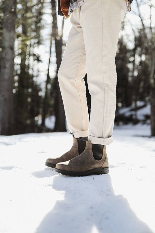 A person wearing light-colored pants and brown Chelsea boots stands on snow in a sunlit forest, with trees and shadows in the background.
