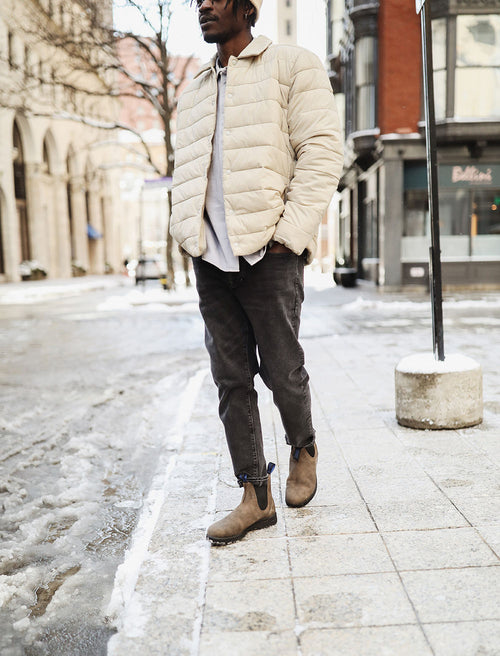 A person wearing a light beige puffer jacket, dark jeans, and brown boots stands on a snowy city sidewalk. The image is cropped above the mouth, showing only the lower face and body.