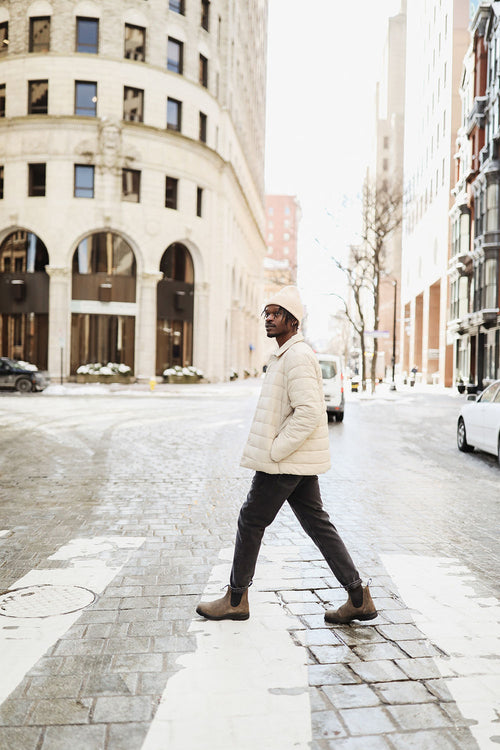 A man wearing a light jacket and beanie walks across a city street at a crosswalk on a bright, sunny day. Tall buildings and a few cars line the street in the background.