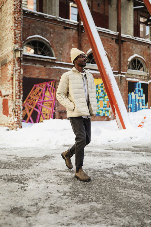 A man wearing a beige puffer jacket, white beanie, dark jeans, and brown boots walks on a snowy street in front of a brick building with colorful graffiti art.