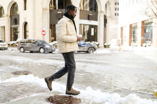 A man wearing a light-colored puffer jacket, black jeans, and brown boots walks across a snowy city street while holding a coffee cup. Cars and buildings are visible in the background.
