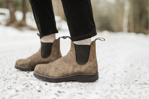 Close-up of a person wearing brown suede Chelsea boots with black elastic panels, standing on a snowy, gravel-covered path. The person is also wearing light-colored socks and black pants.
