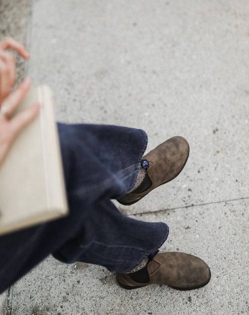A person wearing dark blue jeans, brown shoes, and patterned socks sits on a light-colored concrete surface, holding a closed beige book in their left hand. The image is taken from above.
