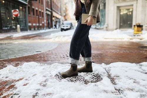 Person standing on a snow-covered street, wearing rolled-up jeans, brown boots, and holding a notebook. Only the lower half of the body is visible; brick buildings and snowy pavement are in the background.