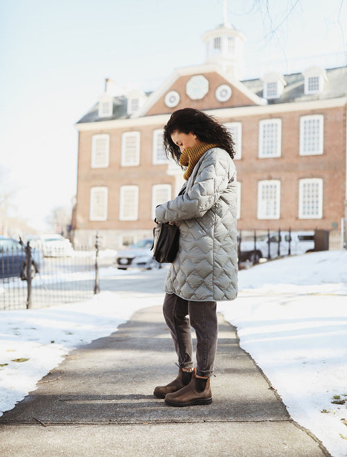 A woman wearing a gray quilted coat, mustard scarf, dark pants, and brown boots stands on a snowy sidewalk, looking into her black bag. A red-brick building with white-trimmed windows is in the background.