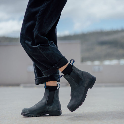 A person wearing black ankle boots and cuffed dark pants stands on a concrete surface outdoors, with a blurred background of buildings and mountains.