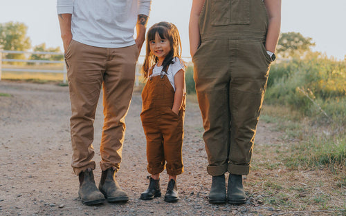 A young girl stands between two adults on a gravel path, all wearing brown pants and boots. The adults faces are not visible. The girl smiles, and the scene is outdoors in warm, golden sunlight.