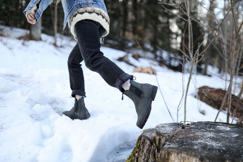 A person wearing black boots, dark pants, and a denim jacket steps onto a snow-covered tree stump in a snowy forest. Only the lower half of their body is visible.