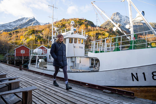 A person in a coat and hat walks along a wooden dock next to a white boat, with snowy mountains and colorful autumn trees in the background. Red cabins are visible on the hillside.