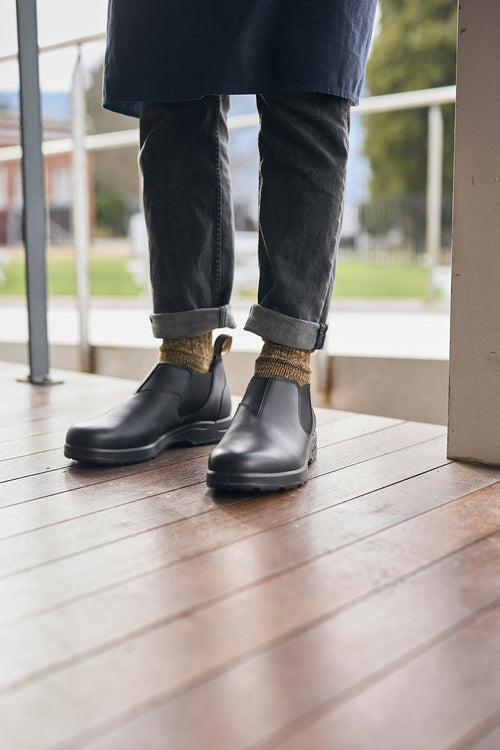 A person wearing rolled-up dark jeans, brown patterned socks, and black slip-on leather boots stands on a wooden floor near a railing with a grassy area visible outside.