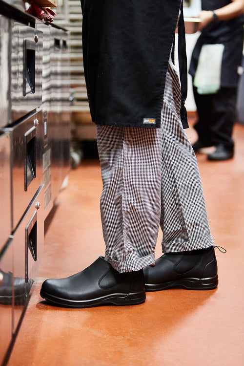 A person wearing checkered chef pants, a black apron, and black slip-resistant shoes stands in a commercial kitchen, with another person blurred in the background.