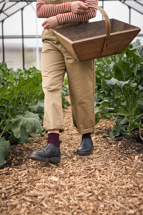 A person wearing tan pants and boots walks through a garden greenhouse, holding a wooden basket by their side. Green leafy plants grow on both sides of a woodchip-covered path.