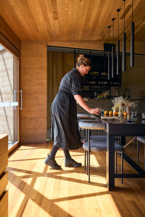A woman in a black dress arranges chairs at a modern dining table set with flowers and small pumpkins in a sunlit room with wooden floors, walls, and ceiling.
