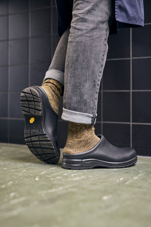 A person wearing gray rolled-up jeans, yellowish wool socks, and black slip-on clogs stands on a greenish floor with one foot lifted, showing the sole. The background features black tiled walls.