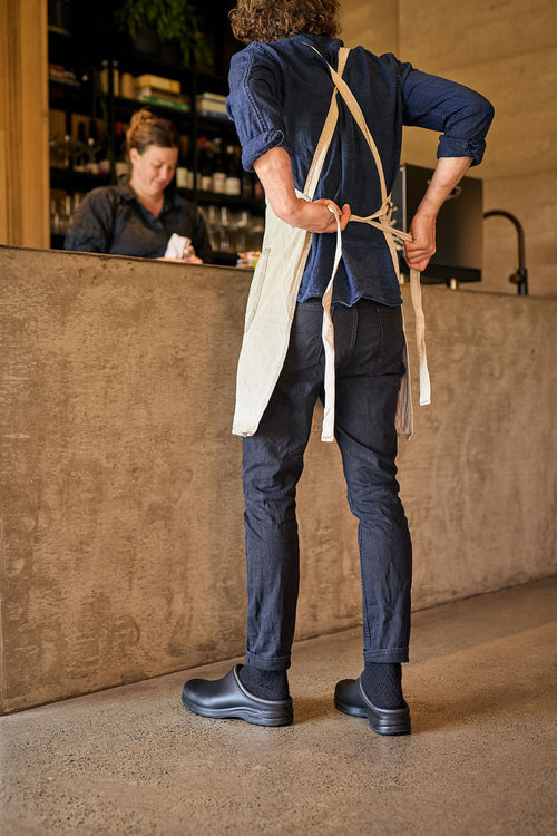 A person in dark clothing and an apron ties the apron behind their back, standing in a modern cafe. Another person is at the counter in the background, partially visible.