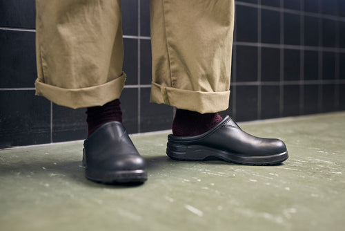 A person wearing rolled-up khaki pants, dark socks, and black slip-on clogs stands on a green floor in front of a black tiled wall.