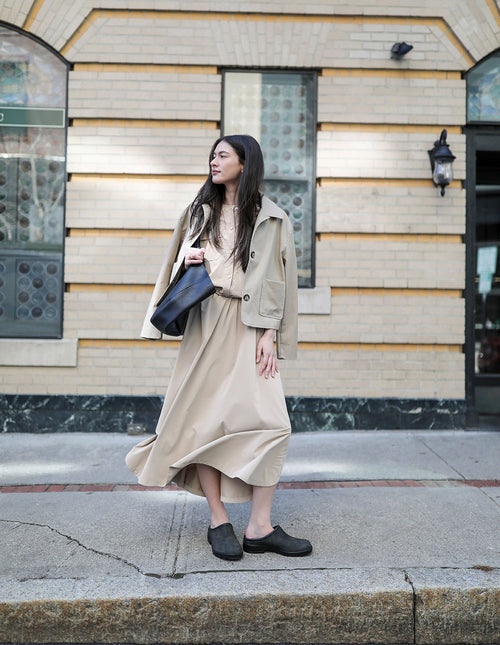 A woman in a beige jacket and long skirt stands on a sidewalk in front of a light brick building, holding a black bag and looking to her left.