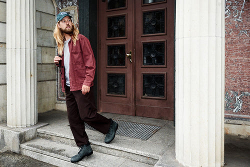 A man with long blond hair and a beard, wearing a green cap, maroon jacket, dark pants, and black boots, walks down stone steps outside a building with large columns and a wooden door.