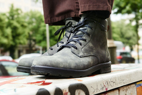A close-up of a person wearing dark gray lace-up boots and brown pants, standing on a graffiti-covered surface outdoors, with blurred trees and cars in the background.