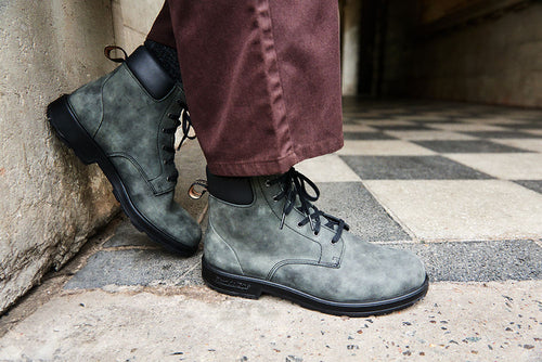 A person wearing green lace-up boots and brown pants stands on a black and white checkered floor next to a concrete wall.