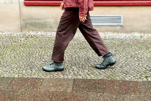 A person wearing brown pants, a reddish jacket, and gray boots walks on a cobblestone sidewalk past a stone wall with a red trim.