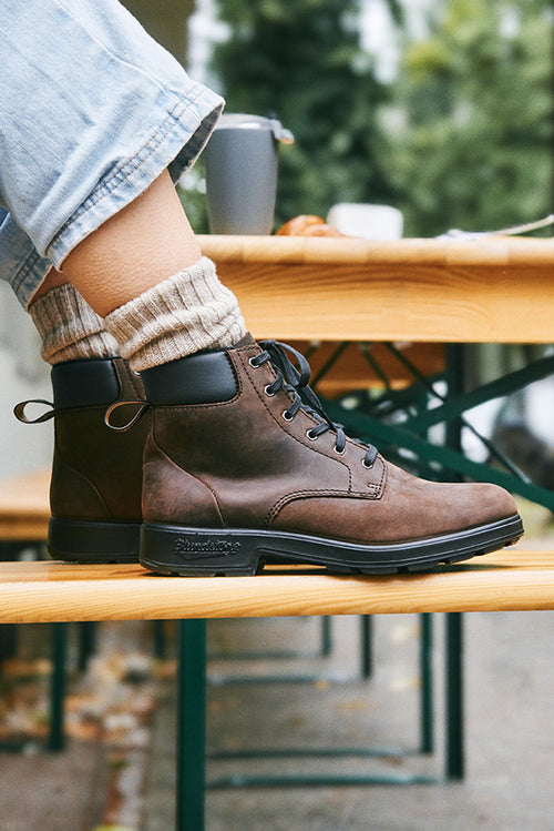 A person wearing light blue jeans, beige socks, and brown lace-up boots sits on a wooden outdoor bench. The scene is casual, with blurred greenery in the background.