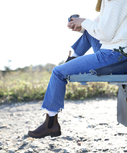 A person wearing blue jeans, brown boots, and a cream sweater sits on the back of a vehicle outdoors, holding a cup, with greenery and rocks in the background.