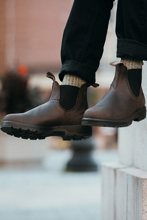 A person wearing dark brown Chelsea boots, gray wool socks, and black pants stands on a stone step outdoors, with one foot slightly raised. The background is softly blurred.