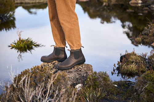 A person wearing brown pants and dark brown boots stands on a rock surrounded by water and vegetation in an outdoor setting.