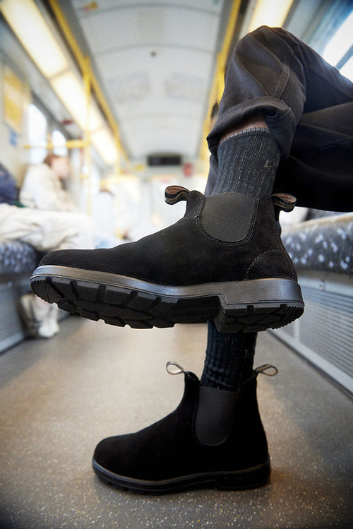 A close-up of a person wearing black Chelsea boots with thick soles, dark socks, and brown pants, sitting on public transport with their legs crossed. The train interior is slightly blurred in the background.