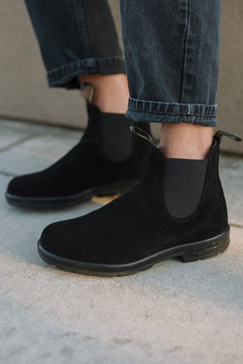Person wearing black suede Chelsea boots with elastic side panels and black jeans with rolled-up cuffs, standing on a light-colored pavement.