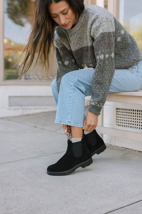 A woman in a gray patterned sweater and light blue jeans sits on a bench, bending forward to adjust her black ankle boots on a concrete sidewalk.